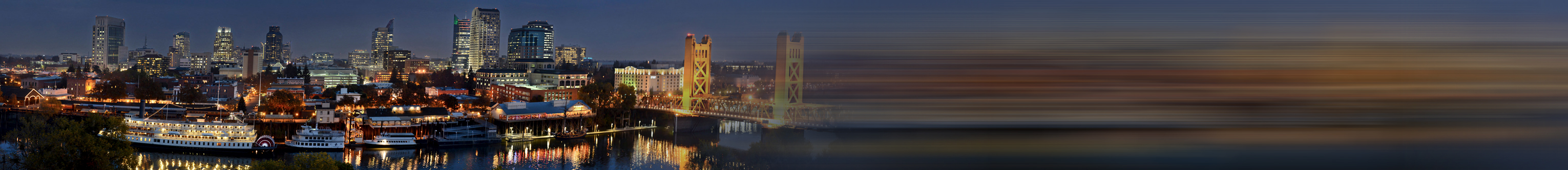 City skyline at dusk with illuminated buildings and a bridge reflected on calm water, blue and amber tones